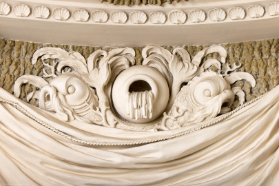 Schloss und Schlossgarten Schwetzingen - Garten - Badhaus - Von innen - Baderaum - Ostwand, oben - Von Westen - Blick auf eine Stuckplastik in der mittleren Mulde des gerafften Vorhangs zwischen den Konsolen; aus einer Urne ergießt sich scheinbar Wasser in das Becken; Urne flankiert von zwei bärtigen Delphinen mit verschlungenen Schwänzen (Stuckateur: Giuseppe Antonio Pozzi (1732–1811)) (Gesamter Bau: Länge x Breite ca. 18 x 14 Meter, Höhe ca. 10 Meter; Architekt: Nicolas de Pigage (1723-1796); Bauzeit: 1768-1772; Gesamtinstandsetzung innen und außen: 1999-2006) (aufgenommen im Oktober 2025, am späten Nachmittag) Schloss und Schlossgarten Schwetzingen - Garten - Badhaus - Von innen - Baderaum - Ostwand, oben - Von Westen - Blick auf eine Stuckplastik in der mittleren Mulde des gerafften Vorhangs zwischen den Konsolen; aus einer Urne ergießt sich scheinbar Wasser in das Becken; Urne flankiert von zwei bärtigen Delphinen mit verschlungenen Schwänzen (Stuckateur: Giuseppe Antonio Pozzi (1732–1811)) (Gesamter Bau: Länge x Breite ca. 18 x 14 Meter, Höhe ca. 10 Meter; Architekt: Nicolas de Pigage (1723-1796); Bauzeit: 1768-1772; Gesamtinstandsetzung innen und außen: 1999-2006) (aufgenommen im Oktober 2025, am späten Nachmittag)