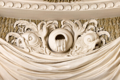 Schloss und Schlossgarten Schwetzingen - Garten - Badhaus - Von innen - Baderaum - Ostwand, oben - Von Westen - Blick auf eine Stuckplastik in der mittleren Mulde des gerafften Vorhangs zwischen den Konsolen; aus einer Urne ergießt sich scheinbar Wasser in das Becken; Urne flankiert von zwei bärtigen Delphinen mit verschlungenen Schwänzen (Stuckateur: Giuseppe Antonio Pozzi (1732–1811)) (Gesamter Bau: Länge x Breite ca. 18 x 14 Meter, Höhe ca. 10 Meter; Architekt: Nicolas de Pigage (1723-1796); Bauzeit: 1768-1772; Gesamtinstandsetzung innen und außen: 1999-2006) (aufgenommen im Oktober 2025, am späten Nachmittag) Schloss und Schlossgarten Schwetzingen - Garten - Badhaus - Von innen - Baderaum - Ostwand, oben - Von Westen - Blick auf eine Stuckplastik in der mittleren Mulde des gerafften Vorhangs zwischen den Konsolen; aus einer Urne ergießt sich scheinbar Wasser in das Becken; Urne flankiert von zwei bärtigen Delphinen mit verschlungenen Schwänzen (Stuckateur: Giuseppe Antonio Pozzi (1732–1811)) (Gesamter Bau: Länge x Breite ca. 18 x 14 Meter, Höhe ca. 10 Meter; Architekt: Nicolas de Pigage (1723-1796); Bauzeit: 1768-1772; Gesamtinstandsetzung innen und außen: 1999-2006) (aufgenommen im Oktober 2025, am späten Nachmittag)