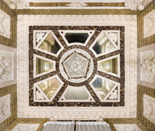 Schloss und Schlossgarten Schwetzingen - Garten - Badhaus - Von innen - Baderaum - Decke - Von unten - Blick von den Stufen ins Wasserbecken nach oben auf die Decke des Raums; von außen nach innen: Kehle, Stuckkonsolen mit birnenförmigen Tropfen, Muscheln, Blütenrosetten, Lorbeergirlande, Streifen Rauputz in Tropfenoptik, dunkle Fläche mit Halbedelsteinen, acht trapezförmige, jeweils mit Stuckrahmen eingefaßte Spiegel, radial angeordnet, in Dreieckskomposition der Eckspiegel Puttenköpfe, in Deckenmitte eine stuckierte Muschel- und Korallenkomposition, kreisförmig umfaßt von einer Lorbeergirlande, darunter die Unterseite der fünfeckigen Laterne (Stuckateur: Giuseppe Antonio Pozzi (1732–1811)) (Gesamter Bau: Länge x Breite ca. 18 x 14 Meter, Höhe ca. 10 Meter; Architekt: Nicolas de Pigage (1723-1796); Bauzeit: 1768-1772; Gesamtinstandsetzung innen und außen: 1999-2006) (aufgenommen im Oktober 2025, am späten Nachmittag) Schloss und Schlossgarten Schwetzingen - Garten - Badhaus - Von innen - Baderaum - Decke - Von unten - Blick von den Stufen ins Wasserbecken nach oben auf die Decke des Raums; von außen nach innen: Kehle, Stuckkonsolen mit birnenförmigen Tropfen, Muscheln, Blütenrosetten, Lorbeergirlande, Streifen Rauputz in Tropfenoptik, dunkle Fläche mit Halbedelsteinen, acht trapezförmige, jeweils mit Stuckrahmen eingefaßte Spiegel, radial angeordnet, in Dreieckskomposition der Eckspiegel Puttenköpfe, in Deckenmitte eine stuckierte Muschel- und Korallenkomposition, kreisförmig umfaßt von einer Lorbeergirlande, darunter die Unterseite der fünfeckigen Laterne (Stuckateur: Giuseppe Antonio Pozzi (1732–1811)) (Gesamter Bau: Länge x Breite ca. 18 x 14 Meter, Höhe ca. 10 Meter; Architekt: Nicolas de Pigage (1723-1796); Bauzeit: 1768-1772; Gesamtinstandsetzung innen und außen: 1999-2006) (aufgenommen im Oktober 2025, am späten Nachmittag)
