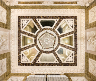 Schloss und Schlossgarten Schwetzingen - Garten - Badhaus - Von innen - Baderaum - Decke - Von unten - Blick von den Stufen ins Wasserbecken nach oben auf die Decke des Raums; von außen nach innen: Kehle, Stuckkonsolen mit birnenförmigen Tropfen, Muscheln, Blütenrosetten, Lorbeergirlande, Streifen Rauputz in Tropfenoptik, dunkle Fläche mit Halbedelsteinen, acht trapezförmige, jeweils mit Stuckrahmen eingefaßte Spiegel, radial angeordnet, in Dreieckskomposition der Eckspiegel Puttenköpfe, in Deckenmitte eine stuckierte Muschel- und Korallenkomposition, kreisförmig umfaßt von einer Lorbeergirlande, darunter die Unterseite der fünfeckigen Laterne (Stuckateur: Giuseppe Antonio Pozzi (1732–1811)) (Gesamter Bau: Länge x Breite ca. 18 x 14 Meter, Höhe ca. 10 Meter; Architekt: Nicolas de Pigage (1723-1796); Bauzeit: 1768-1772; Gesamtinstandsetzung innen und außen: 1999-2006) (aufgenommen im Oktober 2025, am späten Nachmittag)