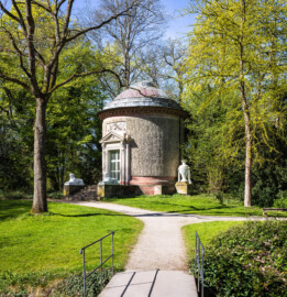 Schloss und Schlossgarten Schwetzingen - Garten - Tempel der Botanik - Von außen - Von Nordost - Blick von der kleinen Brücke über den Leimbachkanal auf den Tempel der Botanik; ein zylindrischer Rundtempel, einem Baumstumpf mit Eichenrinde ähnlich, mit siebenstufiger Treppe, zwei Sphinxen, einem Portal mit Holzlamellen-Flügeltür, Dreiecksgiebel, darin ein geflügelter weiblicher Kopf, darunter eine Inschrift, darüber ein Relief mit zwei Greifen, darüber eine flache Laterne, seitlich zwei steinerne Urnen (Sphinxen Peter Simon Lamine (1738-1817) zugeschrieben; Portal von Franz Conrad Linck (1730-1793); seitliche Urnen von Johann Matthäus van den Branden (1716-1788); Inschrift "Botanicae Silvestri Anno MDCCLXXVIII", deutsch: "Der Waldbotanik im Jahre 1778"); rechts ein Fächerblattbaum (Ginkgobaum / Ginkgo biloba), der am 13. September 2000 zum Gedenken an den 250. Geburtstag von Friedrich Ludwig Sckell (* 13. September 1750, † 24. Februar 1823) nahe dem Tempel der Botanik gepflanzt wurde (Tempel: Durchmesser ca. 7 Meter; Bauzeit: 1777-1780; Architekt: Nicolas de Pigage) (aufgenommen im Mai 2026, am späten Vormittag)