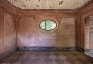 Schloss und Schlossgarten Schwetzingen - Garten - Badhausanlage - Perspektiv - Westlicher Flügel - Von Süden - Blick auf die Nordwand und das Rundfenster des westlichen Flügels; an der Decke die Illusion einer Voliere: ein Gitter, unter dem etliche fliegende Vögel zu sehen sind, darüber ein bewölkter Himmel; Decke und Wände sind mit gemalten rechteckigen Feldern gegliedert, die Ecken tragen Rosettenschmuck; rechts (östlich) die zentrale Halle (Perspektiv: Abmessungen Länge x Breite x Höhe: Laubengang ca. 40 x 3,5 x 4,5 Meter, Bau ca. 16 x 4,5 bis 11 x 6,5 Meter, Bildwand ca. 9 x 1 x 7,5 Meter; Fertigstellung: 1775, Gesamtinstandsetzung: 1988-1991, Restaurierung Laubengang: 2022; Architekt: Nicolas de Pigage) (aufgenommen im April 2026, um die Mittagszeit)