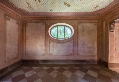 Schloss und Schlossgarten Schwetzingen - Garten - Badhausanlage - Perspektiv - Östlicher Flügel - Von Norden - Blick auf die Südwand und das Rundfenster des östlichen Flügels; an der Decke die Illusion einer Voliere: ein Gitter, unter dem etliche fliegende Vögel zu sehen sind, darüber ein bewölkter Himmel; Decke und Wände sind mit gemalten rechteckigen Feldern gegliedert, die Ecken tragen Rosettenschmuck; rechts (westlich) die zentrale Halle (Perspektiv: Abmessungen Länge x Breite x Höhe: Laubengang ca. 40 x 3,5 x 4,5 Meter, Bau ca. 16 x 4,5 bis 11 x 6,5 Meter, Bildwand ca. 9 x 1 x 7,5 Meter; Fertigstellung: 1775, Gesamtinstandsetzung: 1988-1991, Restaurierung Laubengang: 2022; Architekt: Nicolas de Pigage) (aufgenommen im April 2026, um die Mittagszeit)
