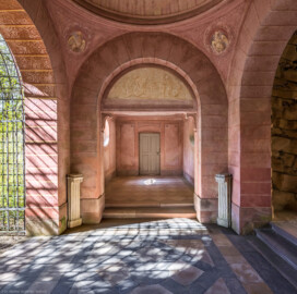 Schloss und Schlossgarten Schwetzingen - Garten - Badhausanlage - Perspektiv - Halle - Von Osten - Blick vom östlichen Seitenflügel nach Westen in die Halle und in den westlichen Seitenflügel; ganz links das Ende des Laubengangs, ganz rechts die Grotte; mittig der Rundbogen mit der Lünette (Darstellung von spielenden Putti als Grisaille), darüber links und rechts zwei Pendentife (Gewölbezwickel) mit Medaillons (Darstellung von Hähnen), ganz oben der Ansatz der Rundkuppel, unten rechts und links zwei Marmorpostamente (ohne Vasen); im Hintergrund die westliche Seitenhalle (Perspektiv: Abmessungen Länge x Breite x Höhe: Laubengang ca. 40 x 3,5 x 4,5 Meter, Bau ca. 16 x 4,5 bis 11 x 6,5 Meter, Bildwand ca. 9 x 1 x 7,5 Meter; Fertigstellung: 1775, Gesamtinstandsetzung: 1988-1991, Restaurierung Laubengang: 2022; Architekt: Nicolas de Pigage) (aufgenommen im April 2026, um die Mittagszeit)