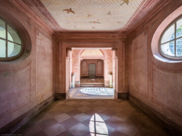 Schloss und Schlossgarten Schwetzingen - Garten - Badhausanlage - Perspektiv - Flügel / Halle - Von Westen - Blick von der Eingangstür im westlichen Flügel nach Osten in die Flügel und die zentrale Halle; zu den Flügeln: an der Decke die Illusion einer Voliere: ein Gitter, unter dem etliche fliegende Vögel zu sehen sind, darüber ein bewölkter Himmel; Decke und Wände sind mit gemalten rechteckigen Feldern gegliedert, die Ecken tragen Rosettenschmuck; zur Halle: zu sehen sind der östliche Rundboden, die östliche Lünette und zwei der vier Marmorpostamente (Perspektiv: Abmessungen Länge x Breite x Höhe: Laubengang ca. 40 x 3,5 x 4,5 Meter, Bau ca. 16 x 4,5 bis 11 x 6,5 Meter, Bildwand ca. 9 x 1 x 7,5 Meter; Fertigstellung: 1775, Gesamtinstandsetzung: 1988-1991, Restaurierung Laubengang: 2022; Architekt: Nicolas de Pigage) (aufgenommen im April 2026, am frühen Nachmittag)