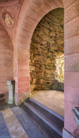 Schloss und Schlossgarten Schwetzingen - Garten - Badhausanlage - Perspektiv - Halle / Grotte - Von Südost - Blick von den Stufen zum östlichen Flügel durch den Rundbogen in die Felsengrotte, geschmückt mit Muscheln, Algen, Halbedelsteinen und Mineralkristallen; links oben das westliche Pendentif mit Medaillon, unten links und rechts zwei Postamente; rechts der Bildmitte die Öffnung zur Vorgrotte, die den Blick auf das Gemälde ("Ende der Welt") freigibt (Perspektiv: Abmessungen Länge x Breite x Höhe: Laubengang ca. 40 x 3,5 x 4,5 Meter, Bau ca. 16 x 4,5 bis 11 x 6,5 Meter, Bildwand ca. 9 x 1 x 7,5 Meter; Fertigstellung: 1775, Gesamtinstandsetzung: 1988-1991, Restaurierung Laubengang: 2022; Architekt: Nicolas de Pigage) (aufgenommen im April 2026, am Nachmittag)