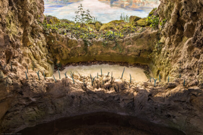 Schloss und Schlossgarten Schwetzingen - Garten - Badhausanlage - Perspektiv - Vorgrotte - Von Süden - Blick von der Mitte der Grotte auf den unteren Teil der Vorgrotte mit dem Wasserbecken und auf das Gemälde "Das Ende der Welt" an der gewölbten Bildwand; Vorgrotte angebaut mit Steinen aus Tuff und Gips, (ehemals) mit Wasser von oben berieselt, unten ein kleines Wasserbecken aus Tuffstein, unten vorne und hinten eiserne Schilfblätter  (Perspektiv: Abmessungen Länge x Breite x Höhe: Laubengang ca. 40 x 3,5 x 4,5 Meter, Bau ca. 16 x 4,5 bis 11 x 6,5 Meter, Bildwand ca. 9 x 1 x 7,5 Meter; Fertigstellung: 1775, Gesamtinstandsetzung: 1988-1991, Restaurierung Laubengang: 2022; Architekt: Nicolas de Pigage) (aufgenommen im April 2026, am frühen Nachmittag)