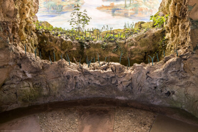 Schloss und Schlossgarten Schwetzingen - Garten - Badhausanlage - Perspektiv - Vorgrotte - Von Süden - Blick von der Mitte der Grotte auf den unteren Teil der Vorgrotte und auf das Gemälde "Das Ende der Welt" an der gewölbten Bildwand; Vorgrotte angebaut mit Steinen aus Tuff und Gips, (ehemals) mit Wasser von oben berieselt, unten ein kleines Wasserbecken aus Tuffstein, unten vorne und hinten eiserne Schilfblätter  (Perspektiv: Abmessungen Länge x Breite x Höhe: Laubengang ca. 40 x 3,5 x 4,5 Meter, Bau ca. 16 x 4,5 bis 11 x 6,5 Meter, Bildwand ca. 9 x 1 x 7,5 Meter; Fertigstellung: 1775, Gesamtinstandsetzung: 1988-1991, Restaurierung Laubengang: 2022; Architekt: Nicolas de Pigage) (aufgenommen im April 2026, am frühen Nachmittag)