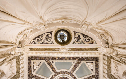 Schloss und Schlossgarten Schwetzingen - Garten - Badhaus - Von innen - Baderaum - Decke - Von unten - Blick vom Wasserbecken nach oben auf den östlichen Teil der Decke; (1) Decke der Wanne in oberer Bildhälfte; von oben nach unten: mehrfach geraffter Stuckvorhang, vier große Konsolen, daran quastenbesetzte Seile befestigt, zwischen Konsolen Stuckplastiken mit Urnen und Delphinen, Fries mit Muscheln, Kreissegment mit drei Feldern, mittig eine Öffnung, mit Tropfenstuck verkleidet, zu einer Laterne zur Entlüftung und Beleuchtung führend, links und rechts davon Felder mit Amethysten und Akanthusranken, darunter ein Feld mit vier Muscheln; (2) Teil der Decke des Raums in unterer Bildhälfte; von außen nach innen: Kehle, Stuckkonsolen mit birnenförmigen Tropfen, Muscheln, Blütenrosetten, Lorbeergirlande, Streifen Rauputz in Tropfenoptik, dunkle Fläche mit Halbedelsteinen, trapezförmige, jeweils mit Stuckrahmen eingefaßte Spiegel, radial angeordnet, in Dreieckskomposition der Eckspiegel Puttenköpfe, in Deckenmitte eine stuckierte Muschel- und Korallenkomposition, kreisförmig umfaßt von einer Lorbeergirlande (Stuckateur: Giuseppe Antonio Pozzi (1732–1811)) (Gesamter Bau: Länge x Breite ca. 18 x 14 Meter, Höhe ca. 10 Meter; Architekt: Nicolas de Pigage (1723-1796); Bauzeit: 1768-1772; Gesamtinstandsetzung innen und außen: 1999-2006) (aufgenommen im Mai 2026, am späten Nachmittag)