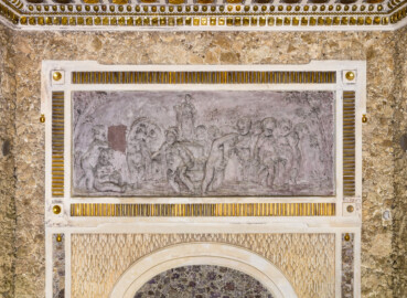 Schloss und Schlossgarten Schwetzingen - Garten - Badhausanlage - Östliches Achathäuschen - Ostwand - Blick auf einen Teil der Hinterwand mit einem Bleirelief; Darstellung eines Erotentanzes um die Statue der Göttin Flora; Flora auf einem Podest stehend, in der Rechten ein Blumenkranz; zehn Putti im Ringeltanz um Flora, teils durch eine Girlande verbunden; links zwei weitere Putti, einander liebkosend; Szene links und rechts eingerahmt durch Bäume (Bildhauer: Franz Conrad Linck (1730-1793), Datierung: um 1770-1775, Material: Blei, Signatur unten rechts (schwach lesbar): "Linck. F", Breite x Höhe: 140 x 57 Zentimeter) (Achathäuschen: Abmessungen Breite x Tiefe x Höhe 3,2 x 3,0 x 6,4 Meter, Innenraum Breite x Tiefe ca. 2 x 2 Meter; Architekt: Nicolas de Pigage, Fertigstellung: um 1775, Instandsetzung: 1991 - 1995) (aufgenommen im Mai 2026, am späten Vormittag) Schloss und Schlossgarten Schwetzingen - Garten - Badhausanlage - Östliches Achathäuschen - Ostwand - Blick auf einen Teil der Hinterwand mit einem Bleirelief; Darstellung eines Erotentanzes um die Statue der Göttin Flora; Flora auf einem Podest stehend, in der Rechten ein Blumenkranz; zehn Putti im Ringeltanz um Flora, teils durch eine Girlande verbunden; links zwei weitere Putti, einander liebkosend; Szene links und rechts eingerahmt durch Bäume (Bildhauer: Franz Conrad Linck (1730-1793), Datierung: um 1770-1775, Material: Blei, Signatur unten rechts (schwach lesbar): "Linck. F", Breite x Höhe: 140 x 57 Zentimeter) (Achathäuschen: Abmessungen Breite x Tiefe x Höhe 3,2 x 3,0 x 6,4 Meter, Innenraum Breite x Tiefe ca. 2 x 2 Meter; Architekt: Nicolas de Pigage, Fertigstellung: um 1775, Instandsetzung: 1991 - 1995) (aufgenommen im Mai 2026, am späten Vormittag)