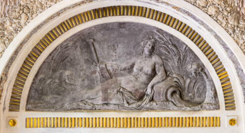 Schloss und Schlossgarten Schwetzingen - Garten - Badhausanlage - Östliches Achathäuschen - Nordwand - Blick auf eine Lünette mit einem Bleirelief an der linken Seitenwand; Darstellung einer Quellennymphe, von Schilfgras eingerahmt; linker Arm gestützt auf Urne, aus der Wasser fließt; rechter Arm ein Ruder haltend; links ein Putto im Schilf (Bildhauer: Franz Conrad Linck (1730-1793), Datierung: um 1770-1775, Material: Blei, Breite x Höhe: 126 x 58 Zentimeter) (Achathäuschen: Abmessungen Breite x Tiefe x Höhe 3,2 x 3,0 x 6,4 Meter, Innenraum Breite x Tiefe ca. 2 x 2 Meter; Architekt: Nicolas de Pigage, Fertigstellung: um 1775, Instandsetzung: 1991 - 1995) (aufgenommen im Mai 2026, am späten Vormittag) Schloss und Schlossgarten Schwetzingen - Garten - Badhausanlage - Östliches Achathäuschen - Nordwand - Blick auf eine Lünette mit einem Bleirelief an der linken Seitenwand; Darstellung einer Quellennymphe, von Schilfgras eingerahmt; linker Arm gestützt auf Urne, aus der Wasser fließt; rechter Arm ein Ruder haltend; links ein Putto im Schilf (Bildhauer: Franz Conrad Linck (1730-1793), Datierung: um 1770-1775, Material: Blei, Breite x Höhe: 126 x 58 Zentimeter) (Achathäuschen: Abmessungen Breite x Tiefe x Höhe 3,2 x 3,0 x 6,4 Meter, Innenraum Breite x Tiefe ca. 2 x 2 Meter; Architekt: Nicolas de Pigage, Fertigstellung: um 1775, Instandsetzung: 1991 - 1995) (aufgenommen im Mai 2026, am späten Vormittag)