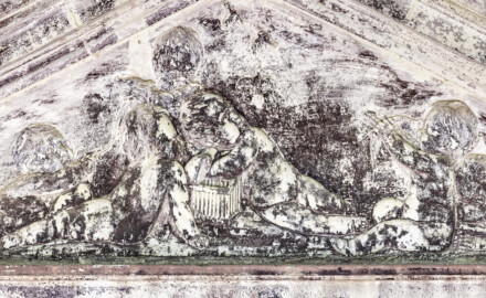 Schloss und Schlossgarten Schwetzingen - Garten - Badhausanlage - Östliches Achathäuschen - Von Westen - Blick vom westlichen Achathäuschen nach Osten auf das (stark verschmutzte) Giebelrelief des östlichen Achathäuschens; Darstellung einer Allegorie des Vogelfangs; unten links der Mitte ein geöffneter Vogelkäfig, aus dem ein Vogel entweicht; rechts des Vogelbauers ein erster Putto, der den Käfig geöffnet hat und dem Vogel nachschaut; links des Käfigs, mit dem Rücken zum Betrachter, ein zweiter Putto, mit beiden Händen auf dem Boden abstützend, ebenfalls dem Vogel nachblickend; hinten ein dritter Putto, einen Vogel in seinen Händen haltend, den ein vierter Putto (ganz links) an einer Schnur gefangen hat; ganz rechts ein fünfter Putto, mit Pfeil und Bogen, auf den entflohenen Vögel zielend; hinter ihm ein schlafender sechster Putto (Bildhauer: Franz Conrad Linck (1730-1793), Datierung: um 1770-1775, Signatur (unten Mitte) "LINCK. F", Material: Sandstein, Breite x Höhe: 140 x 57 Zentimeter) (Achathäuschen: Abmessungen Breite x Tiefe x Höhe 3,2 x 3,0 x 6,4 Meter, Innenraum Breite x Tiefe ca. 2 x 2 Meter; Architekt: Nicolas de Pigage, Fertigstellung: um 1775, Instandsetzung: 1991 - 1995) (aufgenommen im Mai 2026, um die Mittagszeit)