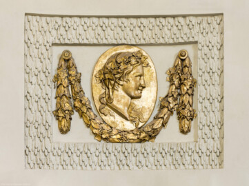 Schloss und Schlossgarten Schwetzingen - Garten - Badhaus - Von innen - Ovalsaal - Vier Jahreszeiten - Von Südost - Nordwestliches Feld mit einem ovalen, bronzierten Medaillon, umfangen von einer Lorbeergirlande, beide umgeben von Tropfenstuck; dargestellt ist ein Frauenkopf im Profil, durch Attribute als Personifikation der Jahreszeit "Winter" erkennbar (Bildhauer: Giuseppe Antonio Pozzi (1732–1811) zugeschrieben) (Gesamter Bau: Länge x Breite ca. 18 x 14 Meter, Höhe ca. 10 Meter; Architekt: Nicolas de Pigage (1723-1796); Bauzeit: 1768-1772; Gesamtinstandsetzung innen und außen: 1999-2006) (aufgenommen im Juli 2026, am frühen Nachmittag)