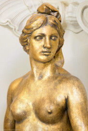 Schloss und Schlossgarten Schwetzingen - Garten - Badhaus - Von innen - Ovalsaal - Nordwestliche Wandnische - Detailansicht des Oberkörpers einer blütenstreuenden Frauenfigur, mit der Rechten einen Gewandbausch mit Blüten raffend, in der linken Hand einen  Blütenbüschel haltend; eine Begleiterin der Aurora, für den Tagesablauf stehend (Bildhauer: Peter Anton von Verschaffelt (1710-1793), Datierung: um 1779, Material: Sandstein, bronziert, Höhe 150 cm) (Gesamter Bau: Länge x Breite ca. 18 x 14 Meter, Höhe ca. 10 Meter; Architekt: Nicolas de Pigage (1723-1796); Bauzeit: 1768-1772; Gesamtinstandsetzung innen und außen: 1999-2006) (aufgenommen im Juli 2026, am späten Nachmittag)