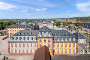 Schloss Bruchsal - Von Außen - Von Oben - Von Süden - Blick vom Kirchturm nach Norden auf das Dach des Finsteren Gangs / Verbindungsgangs (Mitte, ganz vorne), den Kirchenflügel / die Hofkirche (dahinter), den Hauptbau (links, Mitte) sowie auf den Kammerflügel (ganz hinten) (aufgenommen im Mai 2018, am Nachmittag)