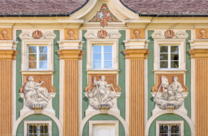 Schloss Bruchsal - Südliche Orangerie / Kavaliersbau - Von Außen - Von Westen - Blick auf die mittleren drei Allegorien bzw. Figuren (v.l.n.r.: vermutlich die "Bildhauerei, die "Architektur" sowie die "Malerei") sowie auf das Allianzwappen im zentralen Giebelfeld (unten: Wappen des Fürstbischofs Franz Christoph von Hutten zum Stolzenberg, links: Wappen des Hochstifts Speyer, rechts: Wappen der Propstei Weißenburg) (ursprünglich gemalt von Giovanni Francesco Marchini, zwischen 1732 und 1737, restauriert von Josef Mariano Kitschker, zwischen 1900 und 1910, erneut restauriert von Klaus Siller (Fassadenmalerei) und Walter Maschke (Allegorien), 1976) (aufgenommen im Oktober 2018, um die Mittagszeit)