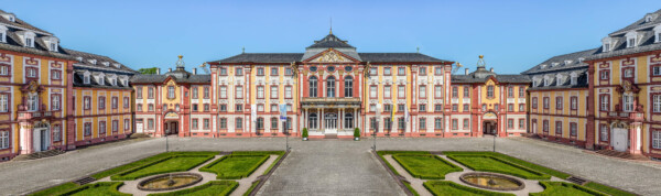 Schloss Bruchsal - Hauptbau / Corps de Logis - Von Außen / Von Osten / Hofseite - Blick vom Balkon des Torwachtgebäudes auf den Ehrenhof, den Hauptbau / das Corps de Logis (Mitte), die Verbindungsbauten (links und rechts) sowie den Kirchenflügel / die Hofkirche / St. Damian und Hugo (ganz links) und den Kammerflügel / das Amtsgericht (ganz rechts) (aufgenommen im Juni 2019, am späten Vormittag)