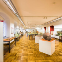 Städtisches Museum Bruchsal - Schloss Bruchsal - Hauptbau / Corps de Logis - Von innen - Südlicher Raum, südliche Hälfte, Bereiche Bronze- / Latène- / Hallstatt- / Römerzeit - Von Osten - Blick vom Eingang in den Raum nach Westen, links Vitrinen mit Fundstücken aus der Bronze- und Römerzeit, in der Mitte vorne Kermaikgefäße aus der Hallstattzeit, dahinter eine Getreide-Handmühle aus der Latènezeit, dahinter Vitrinen mit römischen Fundstücken, rechts ein Eisenbarrenfund mit Doppelpyramidenbarren, dahinter eine Vitrine mit römischen Bronzegefäßen (aufgenommen im Dezember 2020, am frühen Nachmittag)
