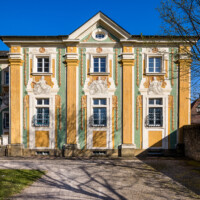 Schloss Bruchsal - Südliche Orangerie / Kavaliersbau - Von Außen / Von Westen - Blick von der südlichsten Ost-West-Wegeachse auf die südliche Westfassade der Orangerie (Fassade der Orangerie ursprünglich gemalt von Giovanni Francesco Marchini, zwischen 1732 und 1737, restauriert von Josef Mariano Kitschker, zwischen 1900 und 1910, erneut restauriert von Klaus Siller (Fassadenmalerei) und Walter Maschke (Allegorien), 1976) (aufgenommen im März 2022, am späten Nachmittag)