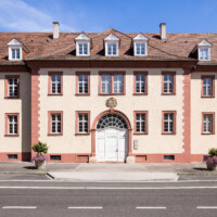 Schloss Bruchsal - Großer Dienerbau / Finanzamt - Mitteltrakt / Mittelrisalit - Von Außen - Von Westen - Blick auf die Westfassade und den Mittelrisalit des Großer Dienerbaus; über dem Durchgang das Landeswappen des Fürstbistums Speyer zur Regierungszeit des Fürstbischofs Kardinal Damian Hugo Philipp von Schönborn-Buchheim (aufgenommen im Juli 2022, am späten Nachmittag)