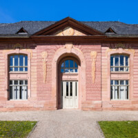 Schloss und Schlossgarten Schwetzingen - Garten - Neue Orangerie - Südseite, Mittelbau - Blick auf den fünfachsigen zweigeschossigen Mittelbau, das rundbogige Portal, den Dreiecksgiebel mit dem Sonnengesicht und den zwei Füllhörnern, die drei Löwenmasken mit Fruchtgehängen sowie die Muscheln mit Füllhörnern über den Fensterbögen (Gesamter Bau: Länge ca. 171 Meter, Breite ca. 10-12 Meter; Bauzeit: 1761-1762; Architekt: Nicolas de Pigage; Putzflächen bemalt in Freskotechnik, täuschen teilweise ein Scheinmauerwerk vor) (aufgenommen im April 2023, am Nachmittag)