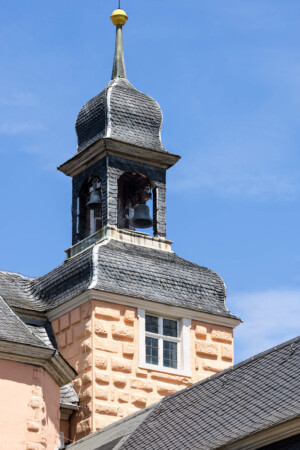 Schloss und Schlossgarten Schwetzingen - Schloss - Mittelbau - Von Südwest - Blick nach oben auf den Südturm des Mittelbaus mit dem Dachreiter, dem Glockenstuhl und den beiden Uhrschlagglocken (Glockengießer: Johann und Andreas Schneidewind / Frankfurt am Main, Gussjahr: 1724; größere / kleinere Glocke: östlich / westlich, Durchmesser 68 cm / 55 cm, Nominal/Schlagton: d'' / f'', Referenznummer (Deutscher Glockenatlas, Bd. 4, Baden, Seite 577): 1891 / 1892; Plattform unter den Glocken: nur ca. 70 x 70 cm; Aufhängung: mittels Holzjochen mit Bandeisenbeschlägen, wohl um 2010 erneuert; Uhrschlaghämmer: elektrisch betrieben und elektronisch gesteuert; Schläge: Stunden- und Viertelstundenschläge) (Mittelbau: Länge Westseite ca. 45 Meter, Breite Süd-/Nordseite ca. 38 Meter; Wiederaufbau / Erweiterung: 1698-1717; Architekten: Johann Adam Breunig, Simon Sarto) (aufgenommen im Mai 2023, am frühen Nachmittag)