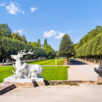 Schloss und Schlossgarten Schwetzingen - Garten - Kreisparterre / Ehemaliges Spiegelbassin / Kugelallee - Hirschgruppe / Hirschbassin - Von Osten - Blick vom Kreisparterre nach Westen auf die rechte (nördliche) Hirschgruppe (samt Hirschbassin) sowie eine Urne (weiter rechts), auf das ehemalige Spiegelbassin (hinter der Hirschgruppe) sowie auf die Kugelallee (noch weiter dahinter) (Bildhauer Hirschgruppe: Peter Anton von Verschaffelt (1710-1793), Datierung: 1766-69; Material Gruppe: gelber Sandstein, Geweih und Hundeschwänze aus Blei, Sockel: roter Sandstein; Abmessungen Gruppe: Höhe ca. 230 cm, Breite ca. 250 cm, Sockel: Höhe 60 cm, Breite 270 cm, Tiefe 270 cm; Bildhauer Urne: Verschaffelt, Datierung: 1773-77; Material Urne: Bleiguß, Sockel aus rotem Sandstein; Urne Höhe 100 cm, Durchmesser ca. 110 cm, Sockel Höhe 65 cm, Breite 70 cm) (aufgenommen im August 2023, am späten Vormittag)