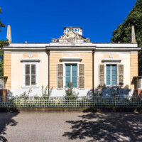 Schloss und Schlossgarten Schwetzingen - Garten - Badhaus - Von aussen - Westseite - Von Westen - Blick auf die Westseite des Badhauses, gegliedert mit drei Achsen, Ecklisenen und mittigem Risalit; in der Mitte oben ein Feld mit Jakobsmuschel und Lorbeerkranz, links oben ein Feld mit Alexander-Medaillon, rechts oben mit Zeus-Medaillon, mittig ganz oben eine ovale Kartusche, von zwei Putti gehalten, mit den Initialen von Carl Theodor ("CT"), darüber der Kurhut mit Kreuz, dahinter das Geländer des Tambours, auf den Ecken oben zwei der acht Obelisken (Gesamter Bau: Länge x Breite ca. 18 x 14 Meter, Höhe ca. 10 Meter; Architekt: Nicolas de Pigage (1723-1796); Bauzeit: 1768-1772; Gesamtinstandsetzung innen und außen: 1999-2006) (aufgenommen im August 2023, am späten Nachmittag)