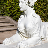 Schloss und Schlossgarten Schwetzingen - Garten - Naturtheater - Parterre - Nordseite, östliche Sphynx - Von Südost - Blick auf eine der sechs Sphyngen (als Allegorien der Musik, des Tanzes, der Komödie, der Tragödie und als Wächterinnen) neben der Treppe zum Zuschauerraum, auf einem Postament liegend (Bildhauer: Peter Anton von Verschaffelt (1710-1793), Datierung: 1766-1773; Material: gelber Sandstein, weiß gefasst, Höhe ca. 100 cm, Postament 160 x 63 cm) (aufgenommen im September 2023, am späten Vormittag)