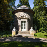 Schloss und Schlossgarten Schwetzingen - Garten - Tempel der Botanik - Von außen - Von Osten - Blick auf den Tempel der Botanik; ein zylindrischer Rundtempel, einem Baumstumpf mit Eichenrinde ähnlich, mit siebenstufiger Treppe, zwei Sphinxen, einem Portal mit Holzlamellen-Flügeltür, Dreiecksgiebel, darin ein geflügelter weiblicher Kopf, darunter eine Inschrift, darüber ein Relief mit zwei Greifen, darüber eine flache Laterne, seitlich zwei steinerne Urnen (Sphinxen Peter Simon Lamine (1738-1817) zugeschrieben; Portal von Franz Conrad Linck (1730-1793); seitliche Urnen von Johann Matthäus van den Branden (1716-1788); Inschrift "Botanicae Silvestri Anno MDCCLXXVIII", deutsch: "Der Waldbotanik im Jahre 1778") (Tempel: Durchmesser ca. 7 Meter; Bauzeit: 1777-1780; Architekt: Nicolas de Pigage) (aufgenommen im September 2023, am frühen Nachmittag)