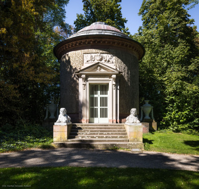 Schloss und Schlossgarten Schwetzingen - Garten - Tempel der Botanik - Von außen - Von Osten - Blick auf den Tempel der Botanik; ein zylindrischer Rundtempel, einem Baumstumpf mit Eichenrinde ähnlich, mit siebenstufiger Treppe, zwei Sphinxen, einem Portal mit Holzlamellen-Flügeltür, Dreiecksgiebel, darin ein geflügelter weiblicher Kopf, darunter eine Inschrift, darüber ein Relief mit zwei Greifen, darüber eine flache Laterne, seitlich zwei steinerne Urnen (Sphinxen Peter Simon Lamine (1738-1817) zugeschrieben; Portal von Franz Conrad Linck (1730-1793); seitliche Urnen von Johann Matthäus van den Branden (1716-1788); Inschrift "Botanicae Silvestri Anno MDCCLXXVIII", deutsch: "Der Waldbotanik im Jahre 1778") (Tempel: Durchmesser ca. 7 Meter; Bauzeit: 1777-1780; Architekt: Nicolas de Pigage) (aufgenommen im September 2023, am frühen Nachmittag)