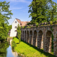 Schloss und Schlossgarten Schwetzingen - Sonstige Bauten - Unteres Wasserwerk - Von Süden - Blick vom westlichen Aquädukt direkt neben dem Römischen Wasserkastell nach Norden auf das Untere Wasserwerk, rechts der nördliche Aquädukt zum Wasserwerk, unten mittig der Leimbach nach Verlassen des Schlossgartens in Fließrichtung Wasserwerk bzw. Norden (Gesamter Bau: Länge x Breite x Höhe: ca. 25 x 13 x 18 Meter; Wasserturm (mit Radkammer, 2 Wasserreservoirs, 3 Geschosse): ca. 13 x 8 Meter, Pumpenhaus (mit Pumpenstube, 2 Geschosse): ca. 14 x 13 Meter, Knochenmühle: ca. 8 x 6 Meter; Architekt: Nicolas de Pigage (1723-1796); Bauzeit: ca. 1762-1765) (aufgenommen im Mai 2024, am frühen Nachmittag)