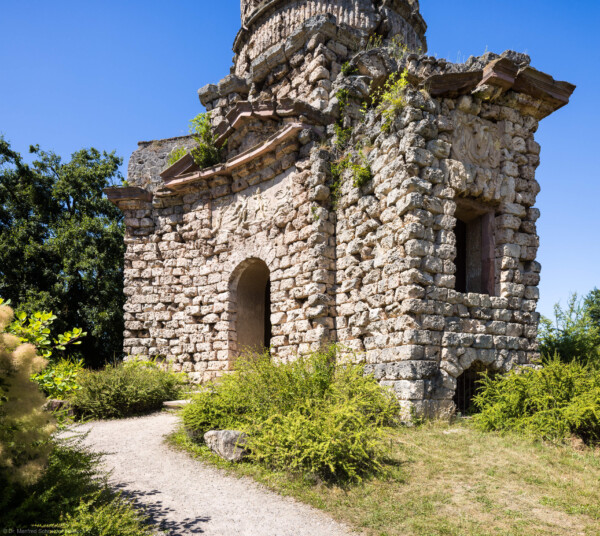 Schloss und Schlossgarten Schwetzingen - Garten - Merkurtempel - Von außen - Von Nordost - Blick auf die breite konkave Ostseite (links) und die schmale Nordseite (rechts) des Merkurtempels, als Ruine gebaut; das Mauerwerk besteht aus Tuffsteinquadern, die Gesimse aus Sandsteinblöcken, die Reliefs aus wetterfestem Marmorstuck; das Feld links zeigt Merkur als Befreier des Prometheus (nach der antiken Sage war aber Herakles / Herkules der Befreier des Prometheus), das Feld rechts zeigt ein Bukranion mit einer Girlande über den Hörnern; links der östliche rundbogige Eingang über wenige Treppenstufen, darüber ein Tympanon (darin ehemals geflügelte Hermesköpfe), rechts mittig ein Fenster eines Kabinetts, rechts unten ein Rundbogenfenster zur Beleuchtung des Untergeschosses (Architekt: Nicolas de Pigage (1723-1796); Bauzeit: 1784-1792, Restaurierung: 2008–2013; Abmessungen: Grundfläche des Sechsecks Länge x Breite ca. 14 x 12 Meter, Länge konkave Portalseiten ca. 10 Meter, Tiefe Kabinettseiten ca. 3,5 Meter, Durchmesser Tambour ca. 6,5 Meter, Höhe ca. 25 Meter, 4 Geschosse) (aufgenommen im Juni 2025, am frühen Nachmittag)