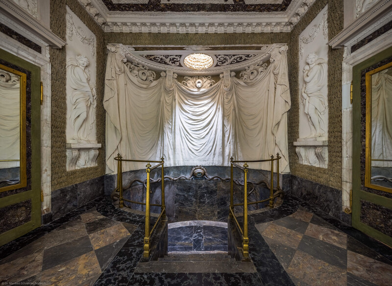 Schloss und Schlossgarten Schwetzingen - Garten - Badhaus - Von innen - Baderaum - Von Westen - Blick nach Osten in das Badezimmer, als Grotte gestaltet; unten in der Mitte sieben Stufen, das ovale Wasserbecken, eingefasst von einem Metallgitter, darüber Wasserspender (zentrale Messingurne und metallene Schlangen), darüber ein großer Stuckvorhang, darüber eine runde Öffnung zur Beleuchtung und Belüftung, ganz links eine Tür nach außen, ganz rechts eine Tür zum westlichen Vorzimmer, links und rechts zwei (von sechs) Wandfelder mit Reliefdarstellungen von Najaden (Nymphen der griechischen Mythologie) (Stuckateur: Giuseppe Antonio Pozzi (1732–1811)) (Gesamter Bau: Länge x Breite ca. 18 x 14 Meter, Höhe ca. 10 Meter; Architekt: Nicolas de Pigage (1723-1796); Bauzeit: 1768-1772; Gesamtinstandsetzung innen und außen: 1999-2006) (aufgenommen im Oktober 2025, am späten Nachmittag)