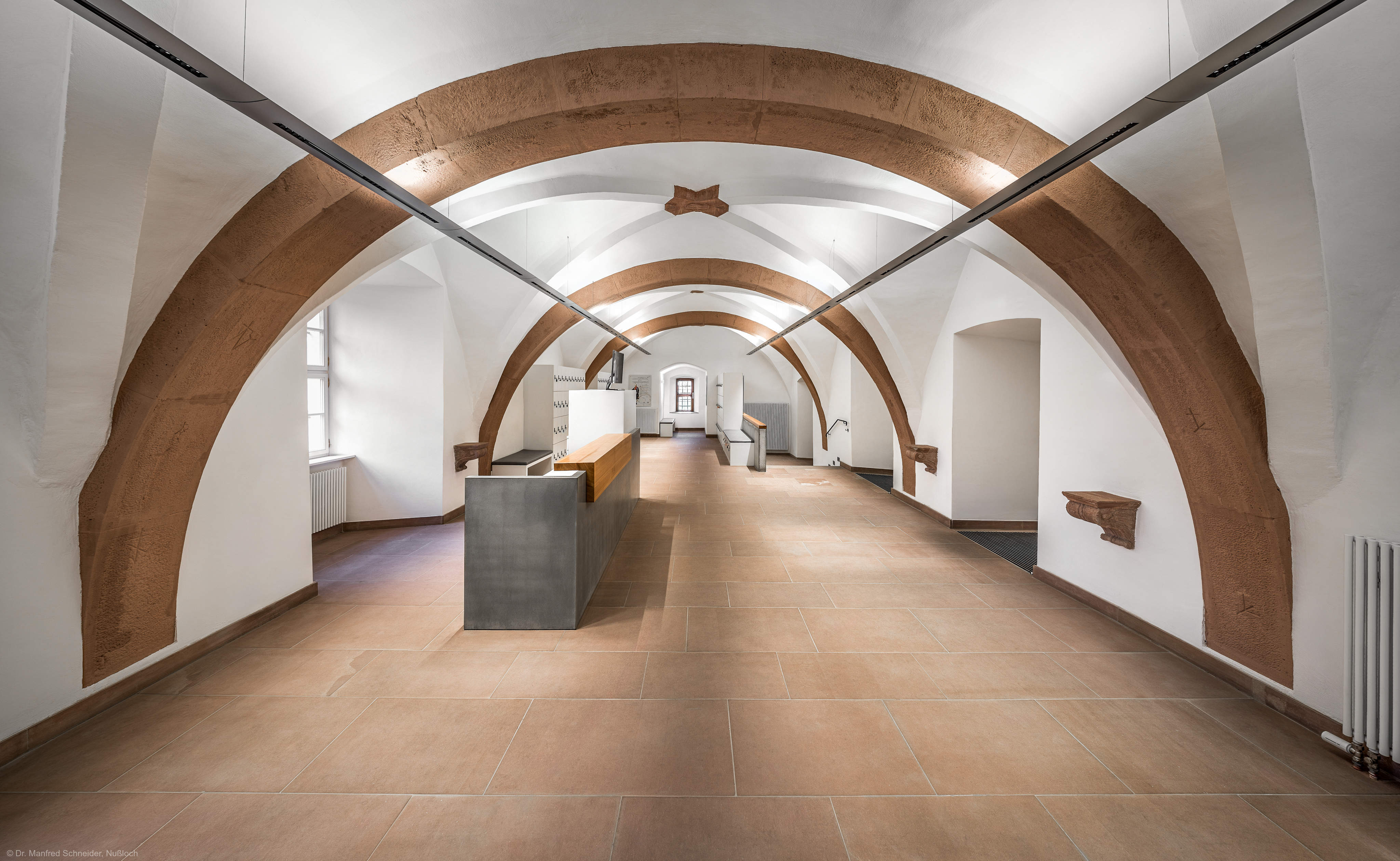 Schloss und Schlossgarten Schwetzingen - Schloss - Mittelbau - Von innen - Erdgeschoss, Nordseite - Besucherzentrum / ehemaliger Hochkeller - Von Westen - Blick von der Westwand des Raums nach Osten in den gewölbten Raum mit 4 Jochen, 3 Gurtbögen und Kreuzgratgewölben; das östliche Ende des Raums und das kleine Fenster sind Teil des Nordturms; in der Mitte rechts zwei Durchgänge zur Vorhalle des Raums, hinten rechts der Durchgang zum nördlichen Treppenhaus; am Ende des Raums hinter der linken (nördlichen Wand) liegt die Schlosskapelle; im 16. Jahrhundert diente der Raum als Hofstube (eine Art Mensa), später als Hochkeller, ab 2016 als Besucherzentrum; der Boden des Raum lag früher 4 Treppenstufen tiefer, der Raum besaß Zwischenwände; ein Gewölbeschlusstein trägt die Jahreszahl 1521 (Mittelbau: Länge Westseite ca. 45 Meter, Breite Süd-/Nordseite ca. 38 Meter; Wiederaufbau / Erweiterung: 1698-1717; Architekt: Johann Adam Breunig) (aufgenommen im November 2025, am frühen Nachmittag)