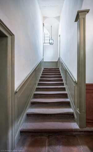 Schloss und Schlossgarten Schwetzingen - Schloss - Mittelbau - Von innen - Nördöstliches Treppenhaus - Von Westen - Blick vom Treppenabsatz im Erdgeschoss nach Osten auf die Treppe und auf den Treppenabsatz zum 1. Obergeschoss; die Tür links führt zur Treppe hinunter in den Besucherraum, die (nicht sichtbare) Tür rechts führt auf den Ehrenhof (Mittelbau: Länge Westseite ca. 45 Meter, Breite Süd-/Nordseite ca. 38 Meter; Wiederaufbau / Erweiterung: 1698-1717; Architekt: Johann Adam Breunig) (aufgenommen im Januar 2026, am frühen Nachmittag)