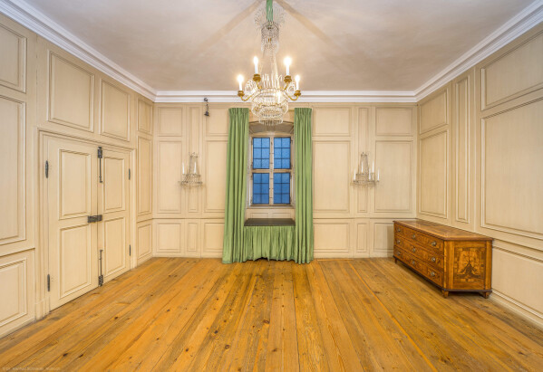 Schloss und Schlossgarten Schwetzingen - Schloss - Mittelbau - Von innen - 2. Obergeschoss - Durchgangszimmer - Nordwand - Von Norden - Blick vom Kamin nach Süden in den Raum und auf eine Kommode; links die Tür zum Flur (aufgenommen im März 2026, am Nachmittag)