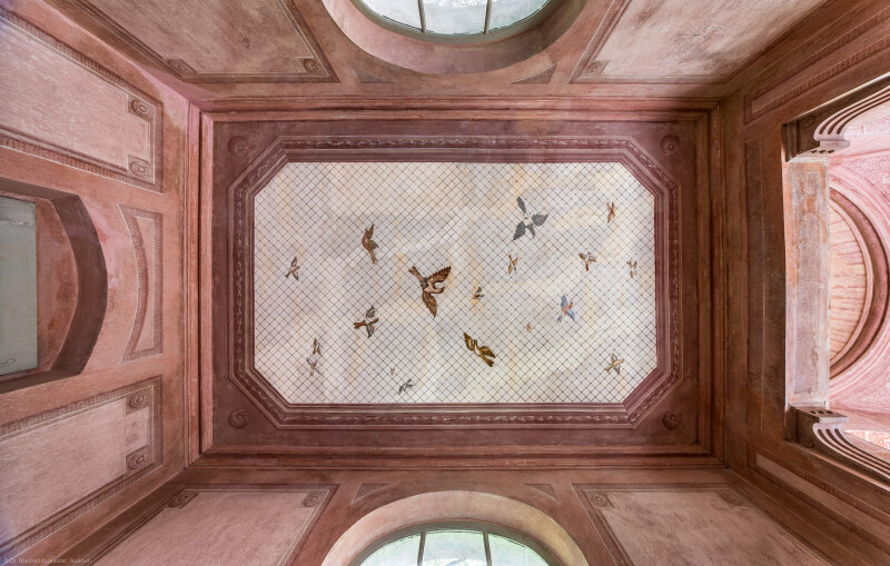Schloss und Schlossgarten Schwetzingen - Garten - Badhausanlage - Perspektiv - Östlicher Flügel - Von unten - Zentraler Blick von der Mitte der östlichen Halle nach oben auf drei Wände und die Decke; an der Decke die Illusion einer Voliere: ein Gitter, unter dem etliche fliegende Vögel zu sehen sind, darüber ein bewölkter Himmel; Decke und Wände sind mit gemalten rechteckigen Feldern gegliedert, die Ecken tragen Rosettenschmuck; links der östliche Eingang, rechts (westlich) die zentrale Halle (Perspektiv: Abmessungen Länge x Breite x Höhe: Laubengang ca. 40 x 3,5 x 4,5 Meter, Bau ca. 16 x 4,5 bis 11 x 6,5 Meter, Bildwand ca. 9 x 1 x 7,5 Meter; Fertigstellung: 1775, Gesamtinstandsetzung: 1988-1991, Restaurierung Laubengang: 2022; Architekt: Nicolas de Pigage) (aufgenommen im April 2026, um die Mittagszeit)
